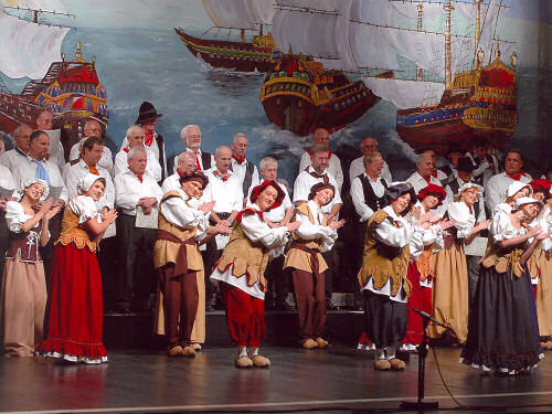 Frauen tanzen den Holzschuhtanz und klatschen dabei in die Hände — dahinter zwei Reihen männlicher Sänger — ganz im Hintergrund das Bühnenbild (riesiges Gemälde mit Schiffen) Foto
